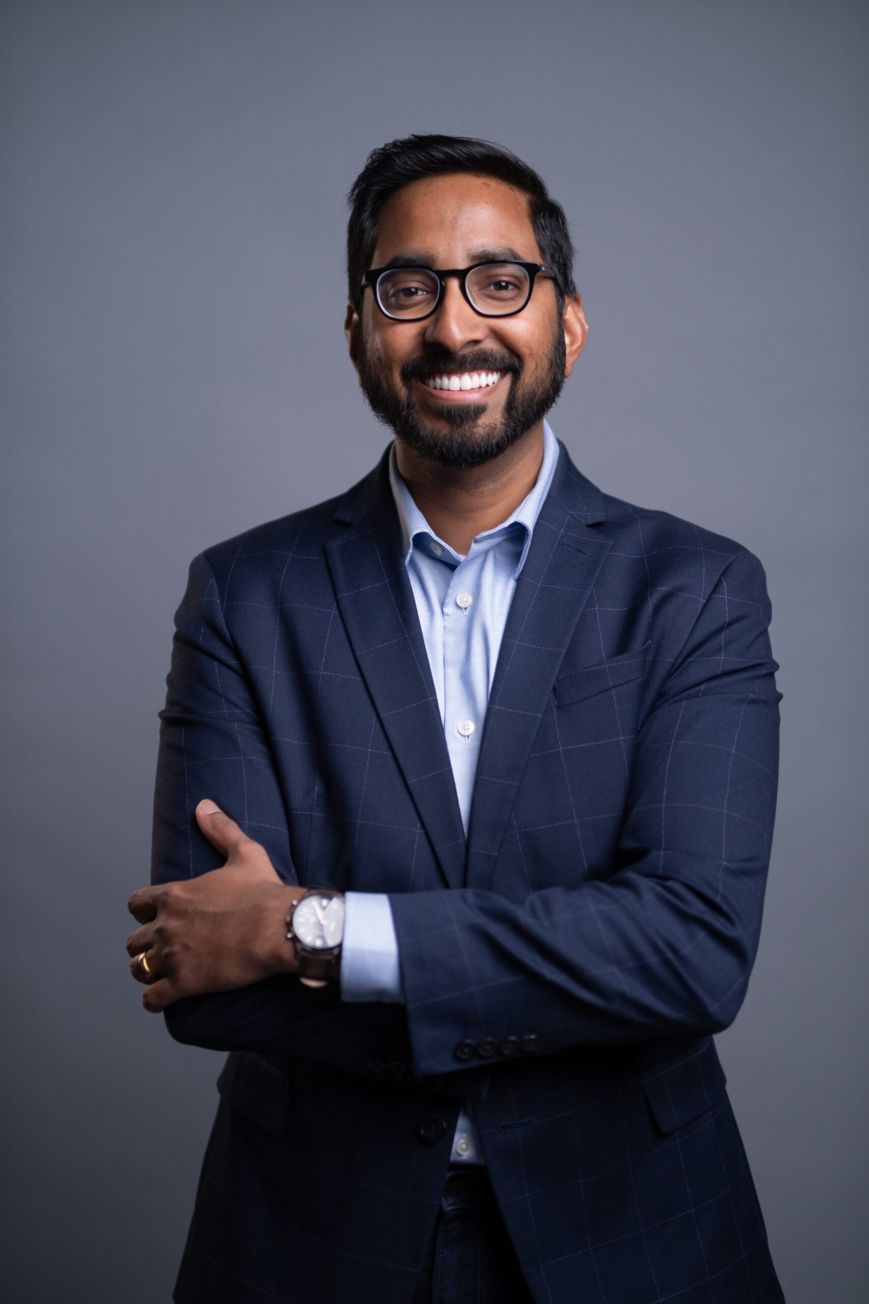 Portrait of a man with short dark hair and glasses, wearing a navy checkered blazer and light blue shirt, smiling against a gray studio background with arms crossed.