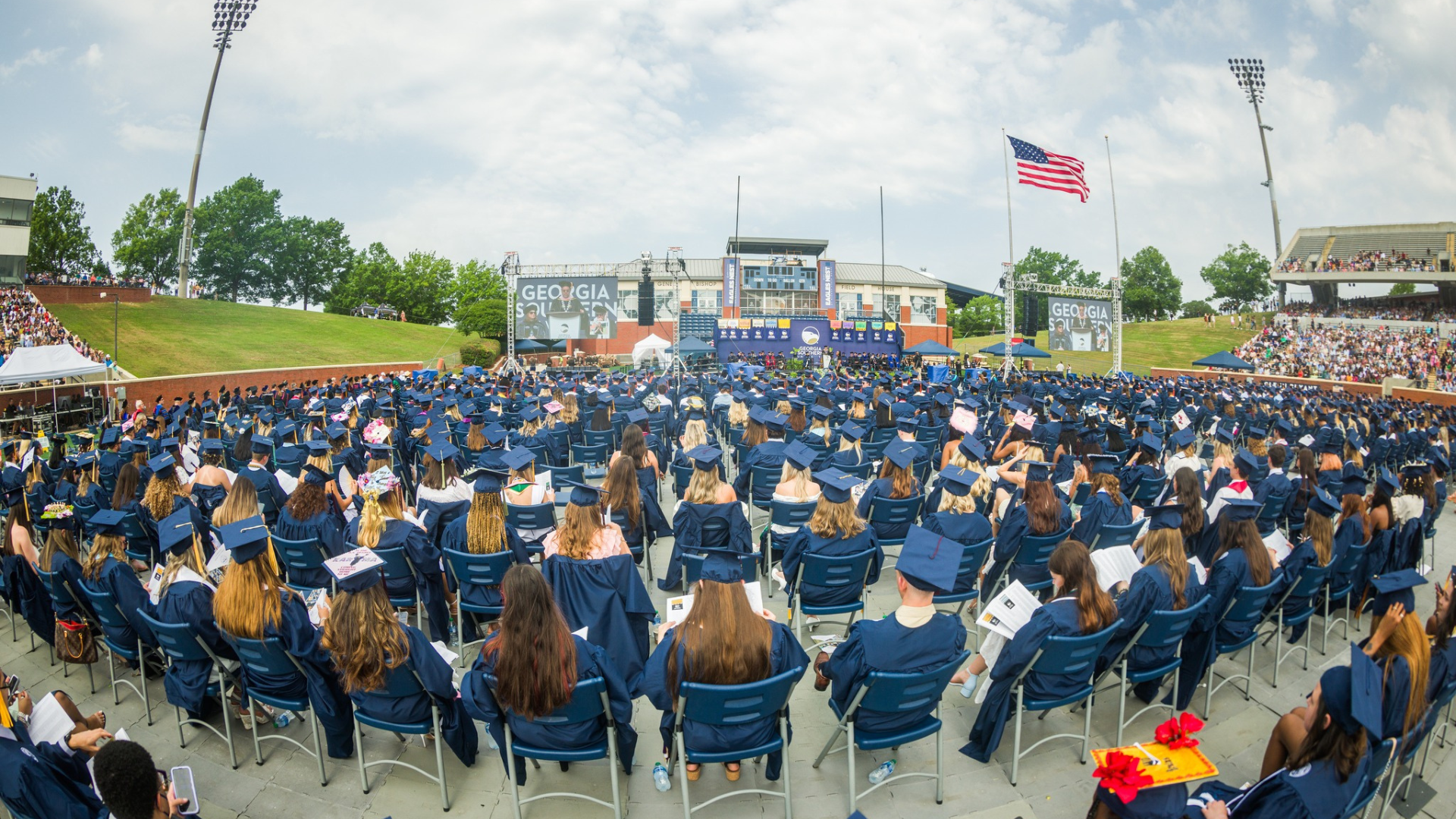 Outdoor graduation ceremony with hundreds of graduates in blue gowns facing a stage at a large stadium; American flag flies nearby.