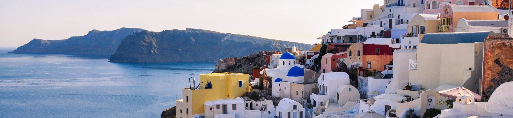 Panoramic view of white-washed buildings with blue domes on a cliffside overlooking the blue Aegean Sea, Santorini.