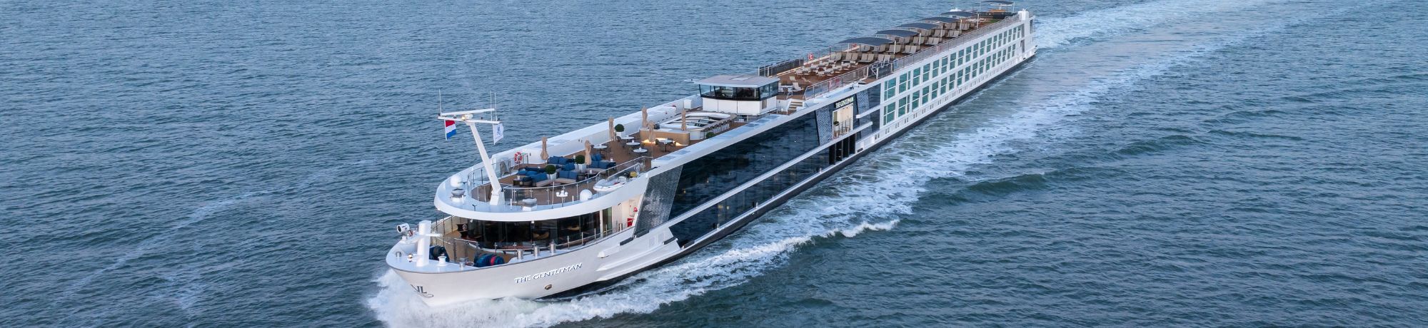 Aerial view of a large white cruise ship sailing on deep blue water, with multiple decks and sun loungers on the top deck.