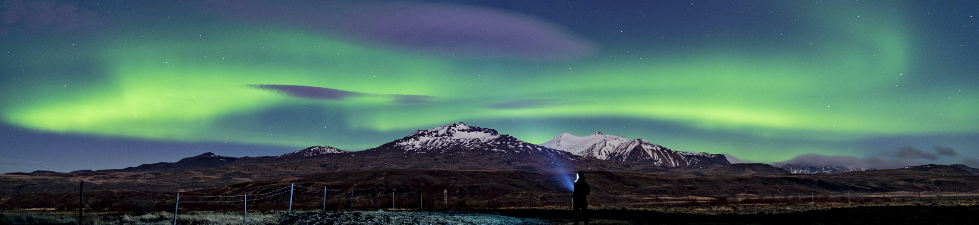 Northern lights glow green across the night sky above snow‑capped mountains; a person in the foreground holds a flashlight.