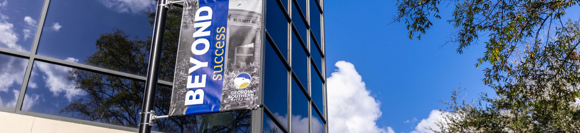 Blue vertical banner reading 'Beyond Success' for Georgia Southern University mounted on a lamp post beside a modern glass building under a bright blue sky.