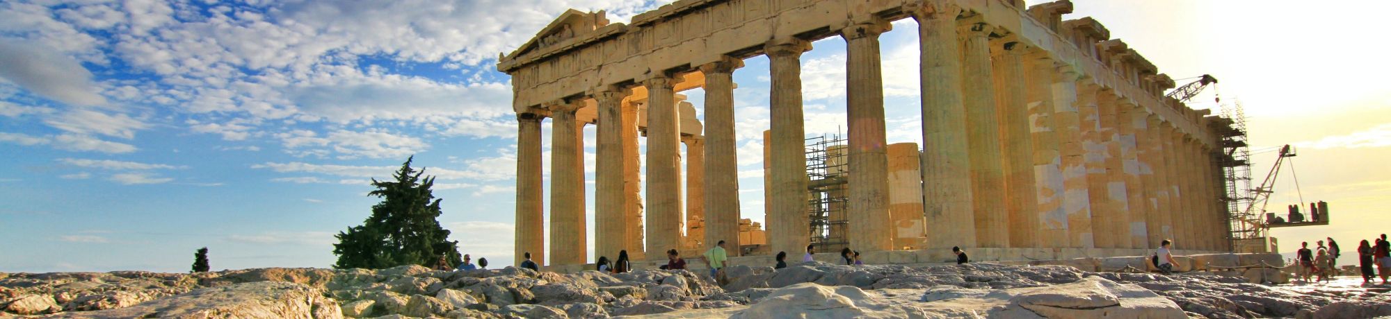 Section of ancient Parthenon-like columns on a rocky foreground with visitors and scaffolding against a bright blue sky.