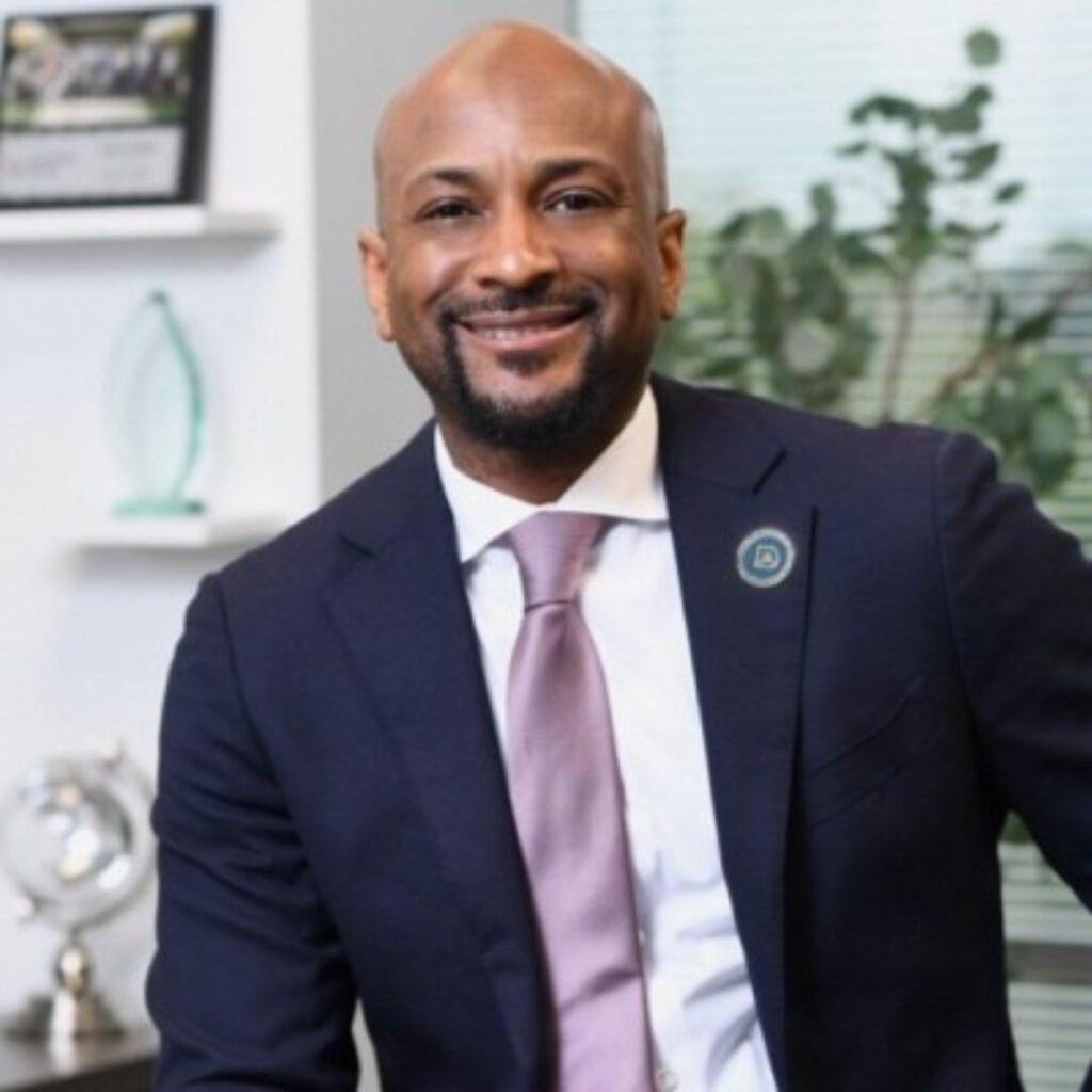 Professional man in a navy suit and pink tie, smiling in an office setting with plants in the background.