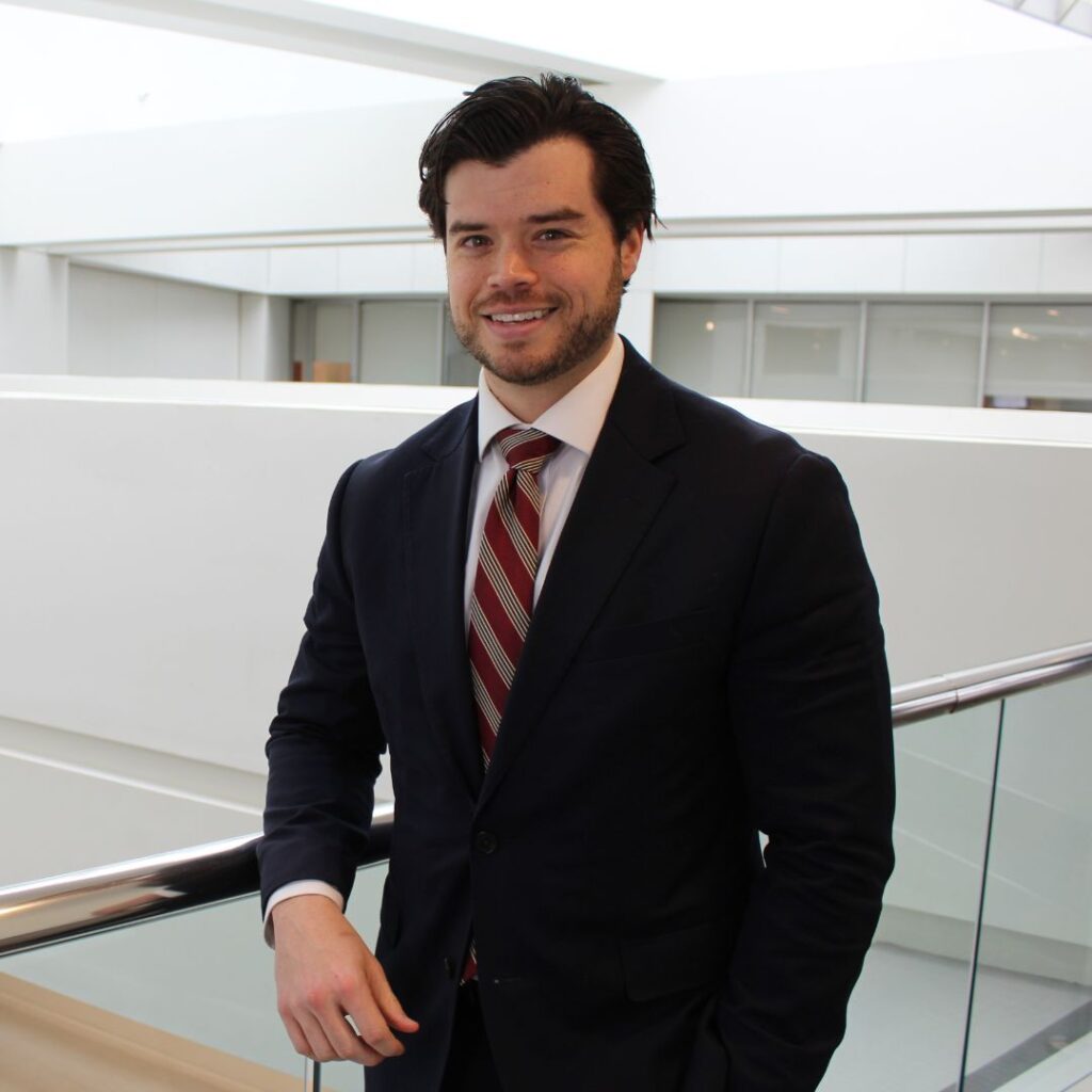 Man in a navy suit with a red striped tie, smiling, leaning on a glass railing in a bright modern office interior.