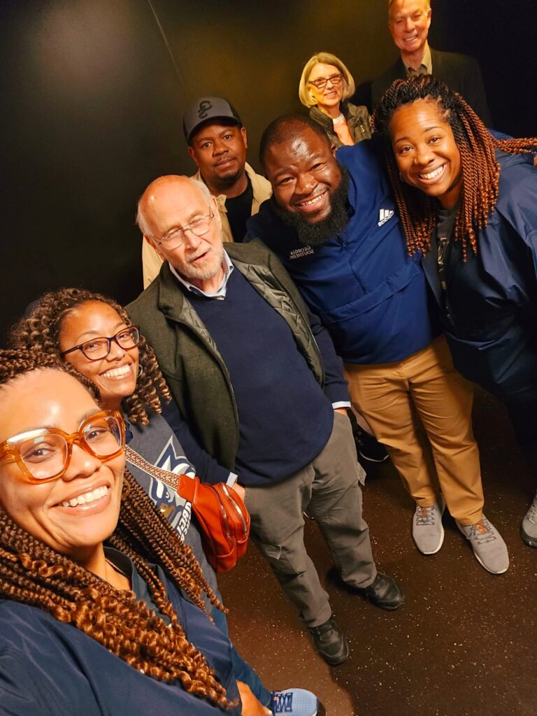 Group of smiling diverse adults posing for a selfie in a dark hallway or elevator. Top of the frame shows people leaning in toward the camera.