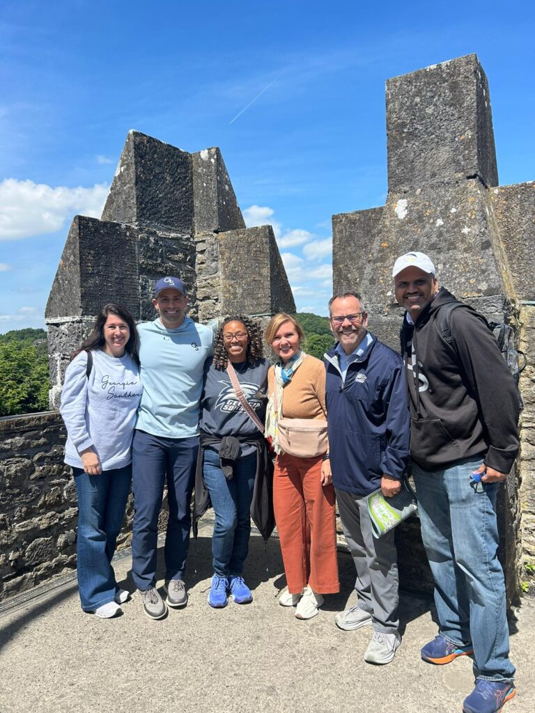 Six adults posing together on a stone castle wall under a bright blue sky on a sunny day.