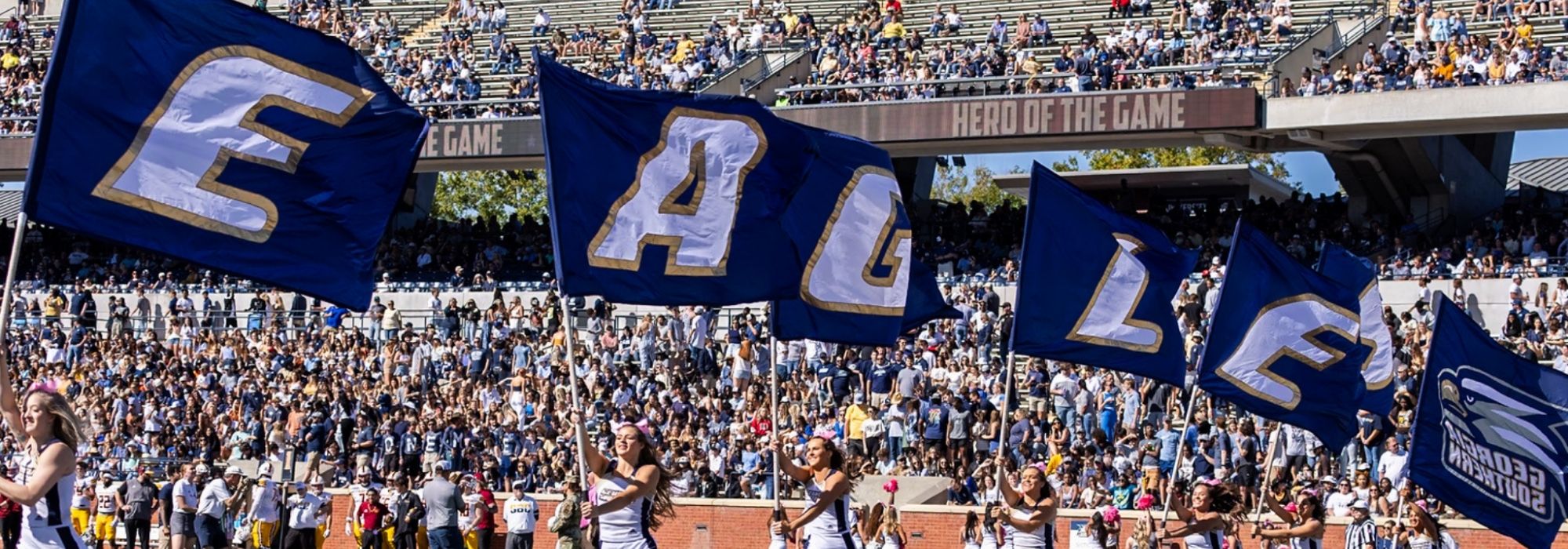 Crowd in a stadium waving blue flags with white letters during a sporting event on a sunny day.