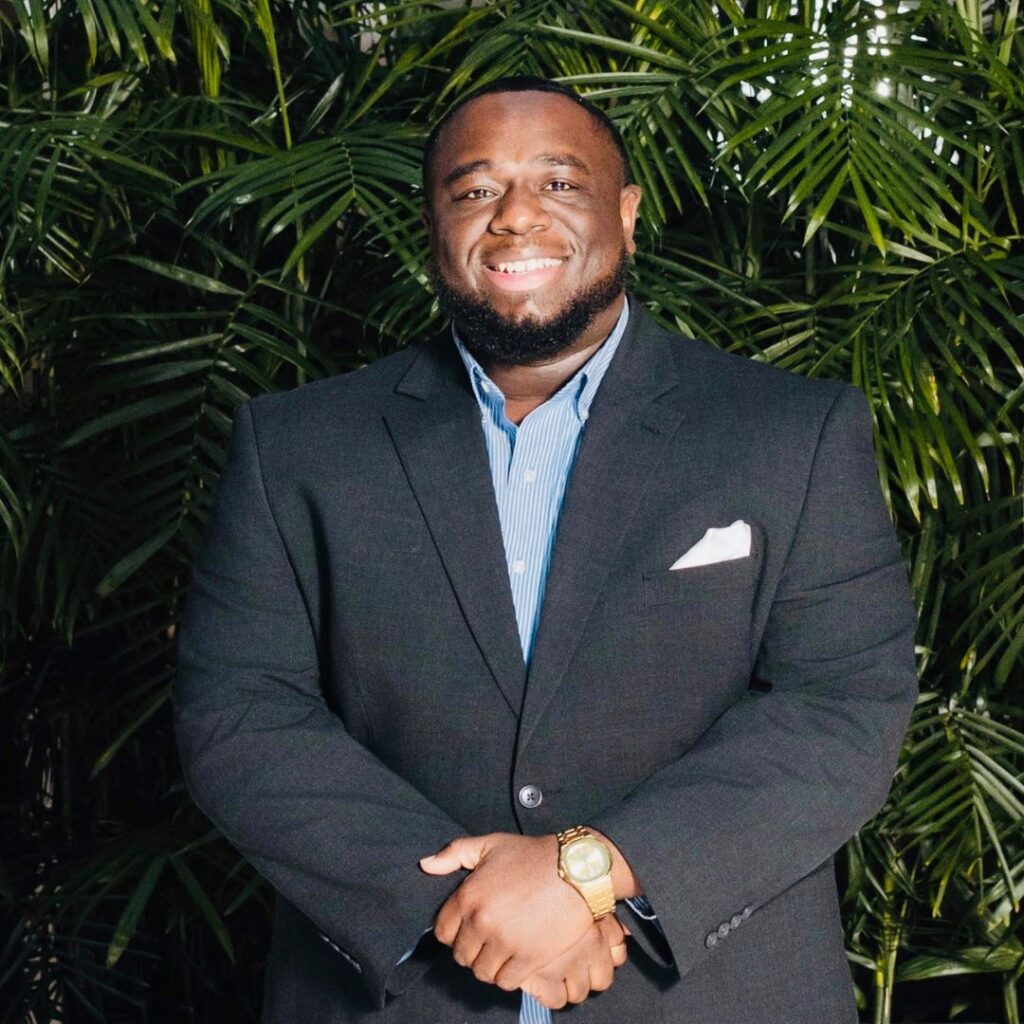 Smiling Black man in a dark suit with a blue striped shirt and a gold watch, standing in front of tropical plants.